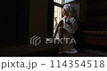Woman in dark room sitting in robe by window. Attractive young girl in bathrobe and with towel on her head is holding cup, looking at window and smiling while sitting on window sill in old hotel room 114354518
