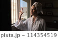 Woman in dark room old hotel standing in robe. Young leady in bathrobe and with towel on her head, looking in window and smiling. Girl has opened the shutters and is standing looking out window 114354519
