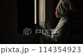 Woman in dark room old hotel standing in robe. Young leady in bathrobe and with towel on her head, looking in window and smiling. Girl has opened the shutters and is standing looking out window 114354923