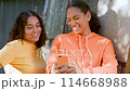 Two beautiful, happy, smiling mixed race African American girls teenagers young women outdoors in the city using a smartphone or cell phone for social media or text messaging 114668988