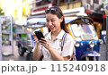 Young Asian woman smiling and using a mobile phone while traveling backpacker in Khaosan Road outdoor market in Bangkok, Thailand 115240918