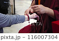 The monk dressed in traditional attire is tying a red string around another person's wrist. It is a religious ceremony that holds special significance in the culture or tradition by the monk. 116007304