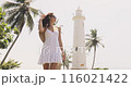 Joyful Woman in White Dress Posing by Tropical Lighthouse 116021422