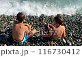 caucasian boys catching waves sitting on pebble sea shore. 116730412