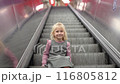 A little girl sits on the steps of an escalator in the subway and goes up. 116805812