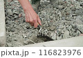 Builder mixing concrete in a black bucket, with the focus on the stirring action. The background consists of gravel, and the person is wearing work pants and sturdy work shoes, indicating a constructi 116823976