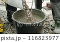 Builder mixing concrete in a black bucket, with the focus on the stirring action. The background consists of gravel, and the person is wearing work pants and sturdy work shoes, indicating a constructi 116823977