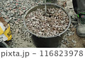 Builder mixing concrete in a black bucket, with the focus on the stirring action. The background consists of gravel, and the person is wearing work pants and sturdy work shoes, indicating a constructi 116823978