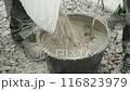 Builder mixing concrete in a black bucket, with the focus on the stirring action. The background consists of gravel, and the person is wearing work pants and sturdy work shoes, indicating a constructi 116823979