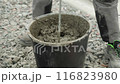 Builder mixing concrete in a black bucket, with the focus on the stirring action. The background consists of gravel, and the person is wearing work pants and sturdy work shoes, indicating a constructi 116823980