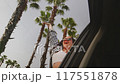 Woman in a car enjoys palm trees of seaside road, leans out of window. The hair is moving in wind 117551878