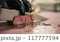Chef in black gloves slices a freshly cooked beef steak with a knife. Cook cuts a hot piece of meat on a kitchen board. Close-up. 117777594