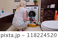 Woman in her 30s sits by a washing machine with a basket of clean laundry in a bright bathroom 118224520