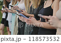 Group of young women are standing in a line outdoors, each looking down at their smartphones. 118573532