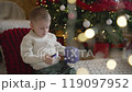 A boy in a jacket examines a Christmas New Year's gift against the backdrop of a decorated room for Christmas. Christmas holidays concept. The boy unpacks a New Year's gift on the background of a 119097952