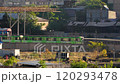 A green train moves through an industrial area with overgrown vegetation and aging structures. 120293478