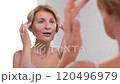 A positive woman in her 50s using moisturizing cream as she touches her face while finger-tipping. Self love and skincare rituals. 120496979