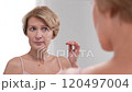 A mature woman is thoughtfully examining her skin in the mirror, confronting the new wrinkles that appear with a mix of emotion and care 120497004