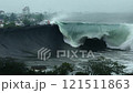 Large wave is crashing into the shore, with a few people in the background 121511863