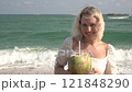 Young blonde woman drinking coconut juice on tropical beach with sea in background, slow motion 121848290