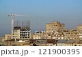 A moving crane works above an unfinished building surrounded by residential structures under a blue sky. 121900395