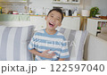 Joyful Asian young boy sitting on couch in a cozy home setting, laughing heartily. His cheerful expression and casual pose convey sense of fun and happiness, surrounded by the warmth and comfort 122597040