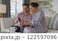 Asian senior couple sitting on couch, happily looking at smartphone screen. They are smiling and sharing moment of connection, showcasing the joy of exploring technology together in cozy home setting 122597096