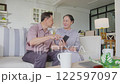 Asian senior couple sitting on couch, happily looking at smartphone screen. They are smiling and sharing moment of connection, showcasing the joy of exploring technology together in cozy home setting 122597097