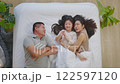 Asian joyful family scene featuring afather, mother, and their young daughter lying comfortably on a bed. The family is enjoying a peaceful and intimate moment, with the child pointing excitedly 122597120