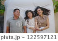 Asian joyful family scene featuring afather, mother, and their young daughter lying comfortably on a bed. The family is enjoying a peaceful and intimate moment, with the child pointing excitedly 122597121