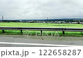 View from a car of a serene countryside village with green fields, houses, and church steeples under an overcast sky. Scenic rural landscape, peaceful horizon, and a calm journey. 122658287