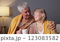 A senior couple sits close together on a sofa, sharing a moment of warmth and affection. They are wrapped in soft blankets, sipping warm beverages, and smiling at each other in a cozy setting 123083582