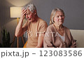 An elderly couple sits apart on a couch in a softly lit living room, showing visible signs of stress 123083586