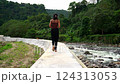 Young girl walking along gray concrete curb along strong mountain river with jungle rainforest background 124313053