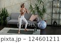 Young blonde woman wearing white sportswear doing squats on a mat in a cozy living room. Gray sofa 124601211