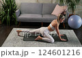 Fit blonde woman working her legs with foam roller on yoga mat in living room. Gray sofa and 124601216