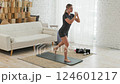 A young fit woman doing yoga on a mat, stepping her leg back. In the background is a white sofa and 124601217