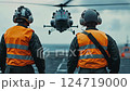 Two crew members of a helicopter on an aircraft carrier, dressed in orange and black uniforms with reflective vests and helmets, look at the camera as a helicopter prepares to land in the background 124719000
