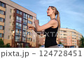 Blonde fitness enthusiast stretching muscular arms, wearing wireless earbuds, preparing for exercise near residential buildings during sunny summer morning 124728451