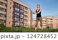 Muscular athlete performing weighted squats on grassy urban landscape, highlighting fitness commitment near residential building setting 124728452