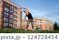 Fitness trainer performing side bends on grassy area near residential complex, highlighting core strengthening and flexibility exercise 124728454