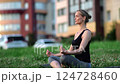 Serene yoga practitioner meditating in lotus pose on urban grass, seeking inner calm amid city noise and movement 124728460
