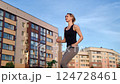 Female athlete executing high-knee running in place, staying fit near residential structures with expansive blue sky surrounding urban workout session 124728461