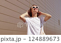Attractive smiling young woman in fashionable sunglasses enjoys sunlights outdoors on warm summer day. Happy stylish girl with short curly hair in good mood. Concept of youth urban lifestyle 124889673