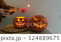 Halloween holidays. Unrecognisable woman kindles pumpkin Jack-O-Lantern by match. Carved two Halloween pumpkins with scary faces and lights on table. Celebration of Day of the dead in autumn at home 124889675