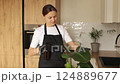 Houseplant care. Young woman in gardening gloves and apron gently sprays water from spray bottle on big green strelitzia leaves iat home kitchen. Watering plants. Sustainable lifestyle, home gardening 124889677