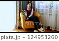 Young woman relaxing in cozy armchair while browsing her phone in bright living room 124953260