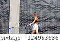 A woman with an empty shopping cart walks along a gray brick wall on a sunny summer day and smiles. Walking towards the supermarket. 124953636