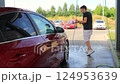 A middle-aged man in a black T-shirt and white shorts washes a red car at a rural self-service car wash in Europe. Washing car with pressure water on the car wash. 124953639