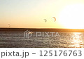 Silhouetted kitesurfers glide across the water during sunset, with vivid golden light reflecting on the sea and kites soaring in the sky 125176763
